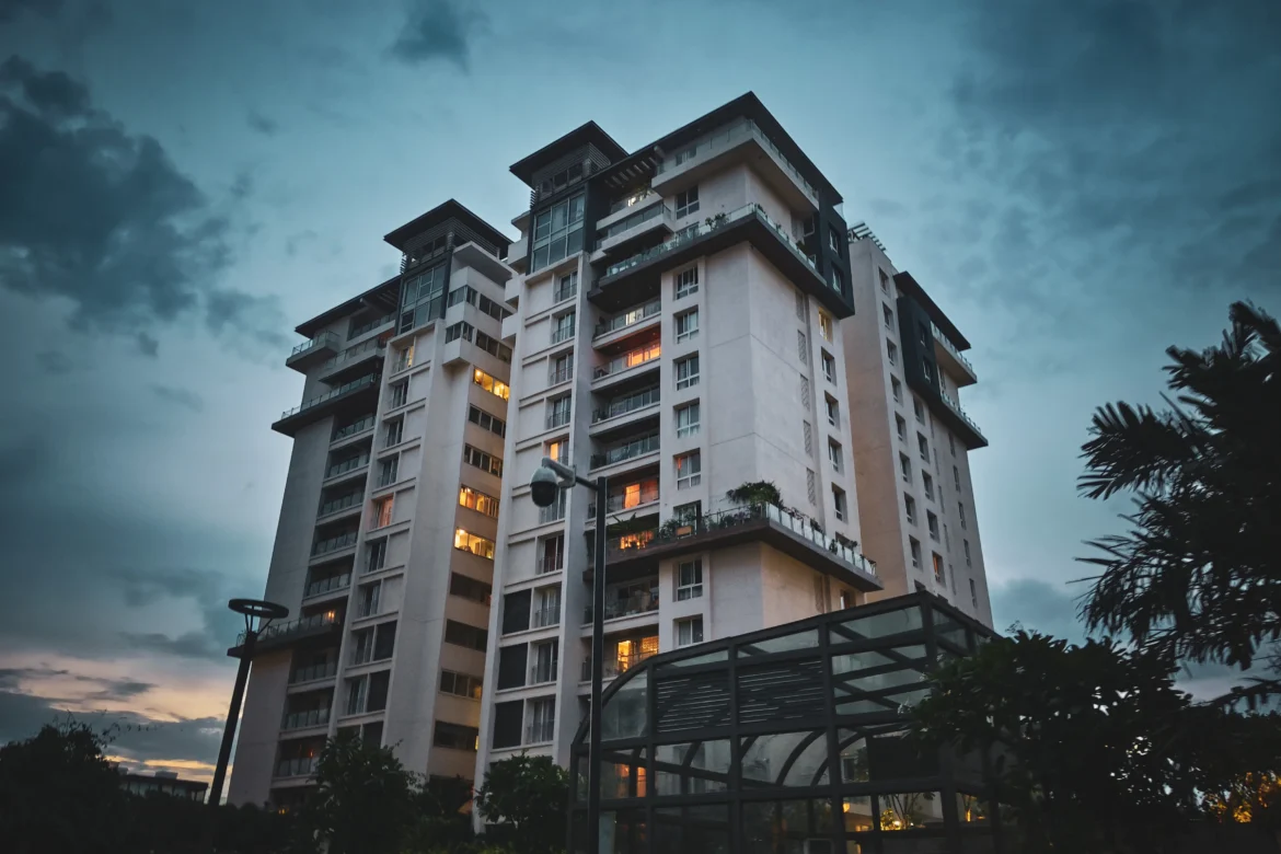 A high-rise modern residential apartment building illuminated with warm lights during dusk, surrounded by trees and urban landscaping under a cloudy evening sky.